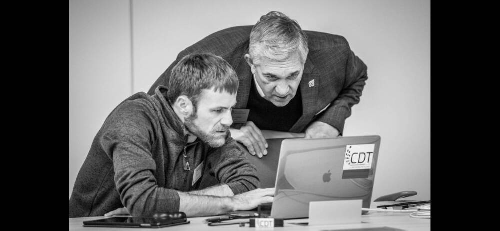 A black and white image of two men one standing and the other sitting both look closely at a laptop